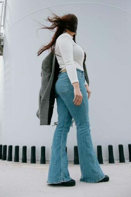 a plus-size woman posing on her side wearing white long sleeves with her jacket on the left arm and bell bottom jeans