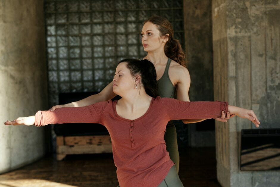 two women; one doing a yoga warrior pose while the other assisting it