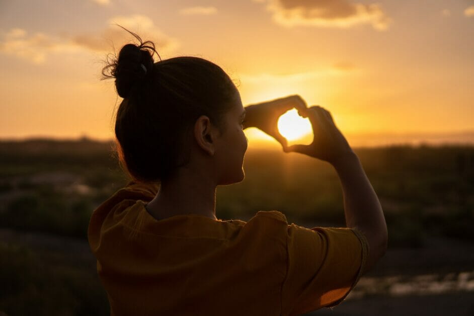 woman forming a heart shape with her hands facing a beautiful sunset