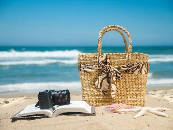 camera at the top of an open notebook besides a summer bag on the sand